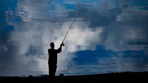 Die Silhouette eines Anglers steht am Ufer des Big Bear Lake und wirft eine Angelschnur ins Wasser. Das Wasser ist blau