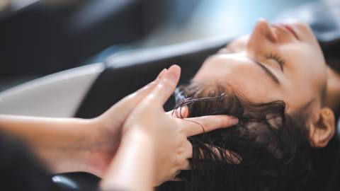 Ein Foto einer Frau, die sich in einem Salon die Haare waschen lässt, während die Finger des Friseurs ihre Kopfhaut massieren.
