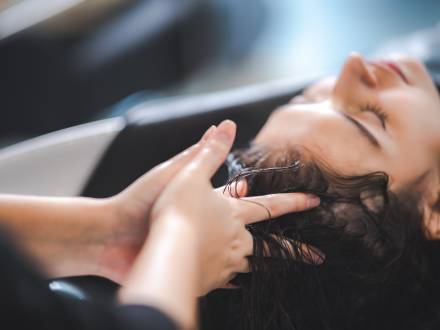 Ein Foto einer Frau, die sich in einem Salon die Haare waschen lässt, während die Finger des Friseurs ihre Kopfhaut massieren.
