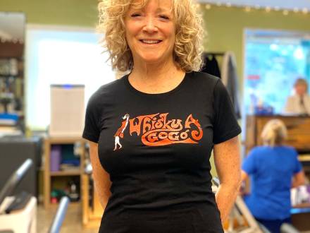 Ein Foto einer Pilates-Lehrerin, die ihre Hände hinter dem Rücken verschränkt und ein einladendes Lächeln zeigt