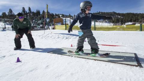 Ein junger Snowboarder rutscht während einer Unterrichtsstunde über ein Rail. Sein Lehrer schaut zu.