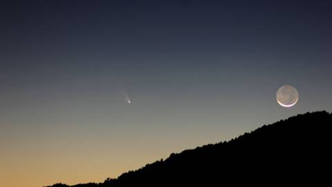 A photo of a silhouetted mountain, the moon is in a waning crescent stage as a comet is passing far away.