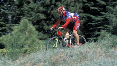 Ein Mountainbiker in einem leuchtend roten Dress fährt auf einem Radweg in Big Bear Lake, Kalifornien. Der üppige, grüne Wald umgibt ihn.