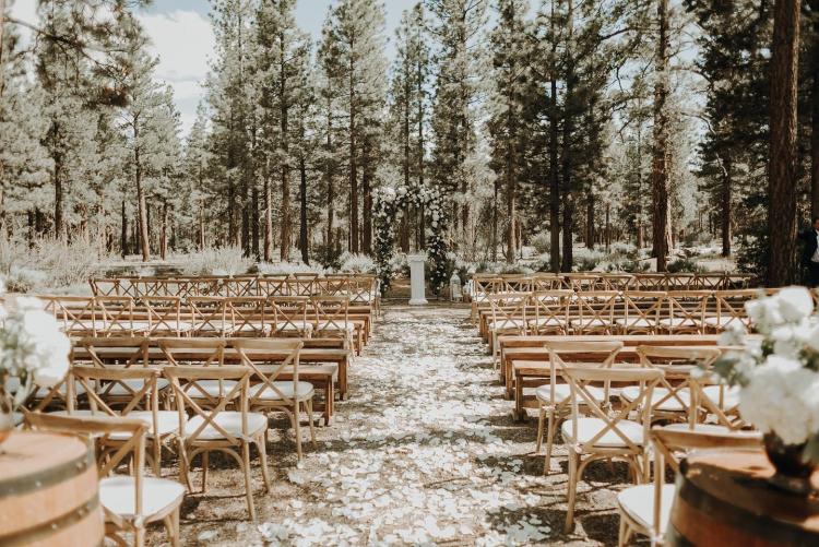 Holzstühle stehen in Reihen an einem bewaldeten Hochzeitsort in Big Bear. Weiße Blumen stehen auf Weinfässern im Vordergrund.