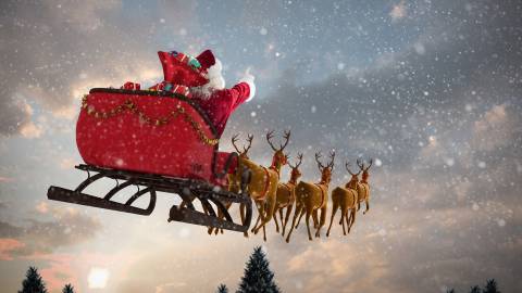 Ein Bild vom Weihnachtsmann, der mit seinem roten Schlitten, gezogen von seinen Rentieren, durch den fallenden Schnee fährt, während die Sonne langsam aufgeht.