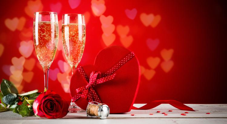 Zwei Champagnerflöten stehen auf einem Tisch mit einer herzförmigen Schachtel mit Schokolade und einer roten Rose in einem Bild zum Thema Valentinstag.