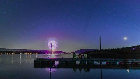 Menschen sitzen auf einem Steg am Big Bear Lake und bewundern das Feuerwerk zum 4. Juli, das sich auf dem See spiegelt.