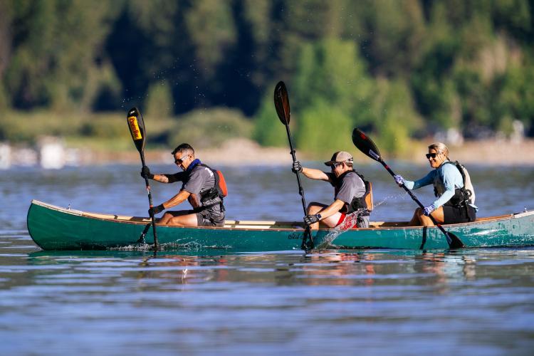 Eine Dreiergruppe auf einem Kajak während des LakeFestes, die gemeinsam mit ihren Paddeln rudert und dabei Wasser nach oben spritzt.