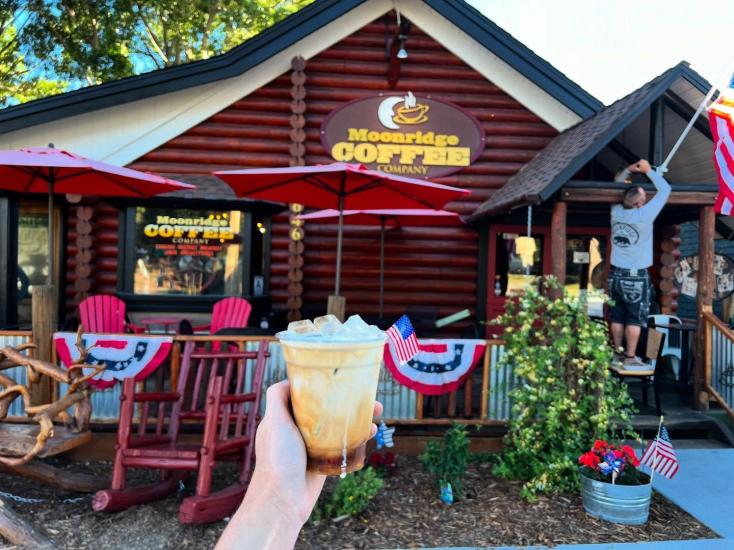 Ein Foto von einer Hand, die einen Eiskaffee vor dem Gebäude von Moonridge Coffee Co. in die Höhe hält.