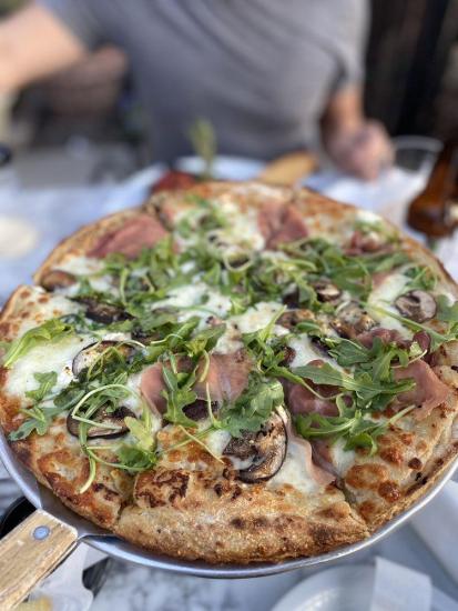 Ein Foto einer Pizza aus der Trattoria La Pergola von oben, mit Rucola, Champignons und geschnittenem Prosciutto.