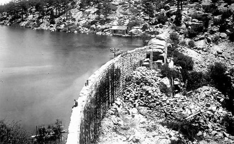 Ein Mann sitzt auf der Kante des ersten, steinernen Big Bear Lake-Damms mit Blick auf den Big Bear Lake vor 1911