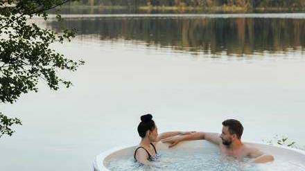 Ein Paar, das sich in einem Whirlpool mit Blick auf den See entspannt, während sich die Bäume in der Ferne sanft im See spiegeln.