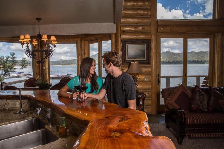 Ein Paar sitzt an einem Holztresen mit Weingläsern in einer Hütte mit Blick auf den Big Bear Lake vor den Fenstern.