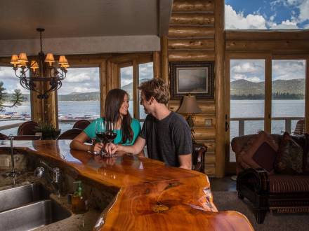 Ein Paar sitzt an einem Holztresen mit Weingläsern in einer Hütte mit Blick auf den Big Bear Lake vor den Fenstern.